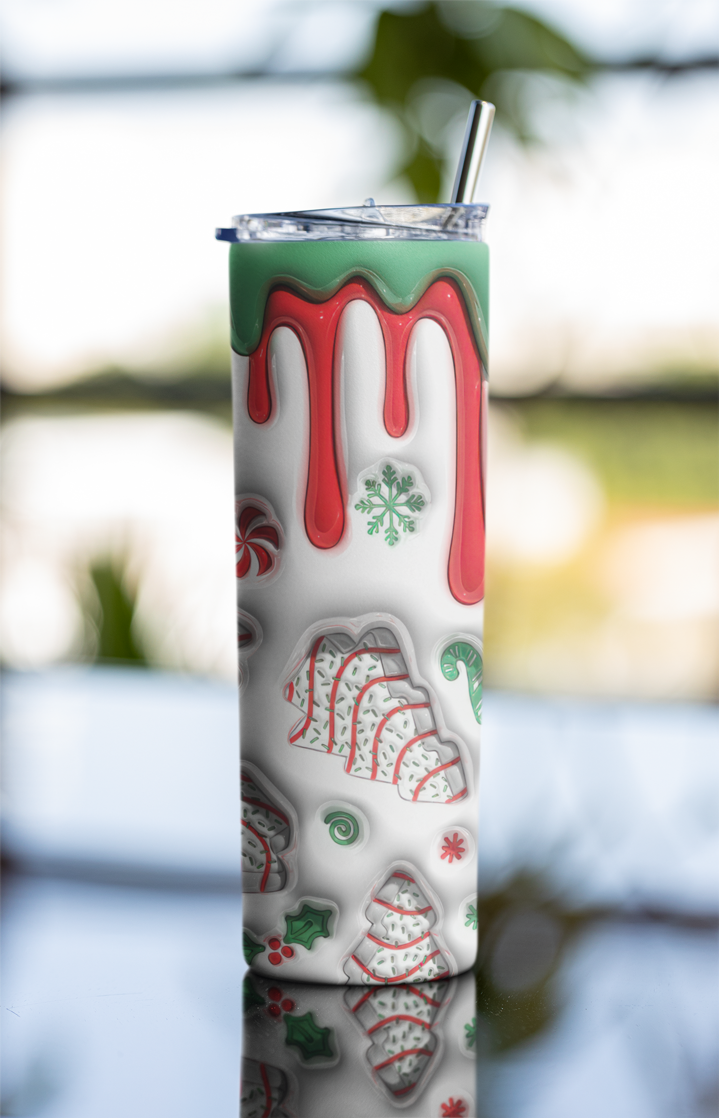 A tall insulated tumbler with a clear lid and metal straw, decorated with a holiday-themed pattern featuring red dripping icing, green snowflakes, peppermint candies, and gingerbread cookies on a white background.