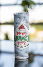 A tall insulated tumbler with a clear lid and metal straw, printed with a Grinch-themed holiday design including the text 'Merry Grinchmas' and snowflake motifs, photographed against a softly blurred outdoor background.