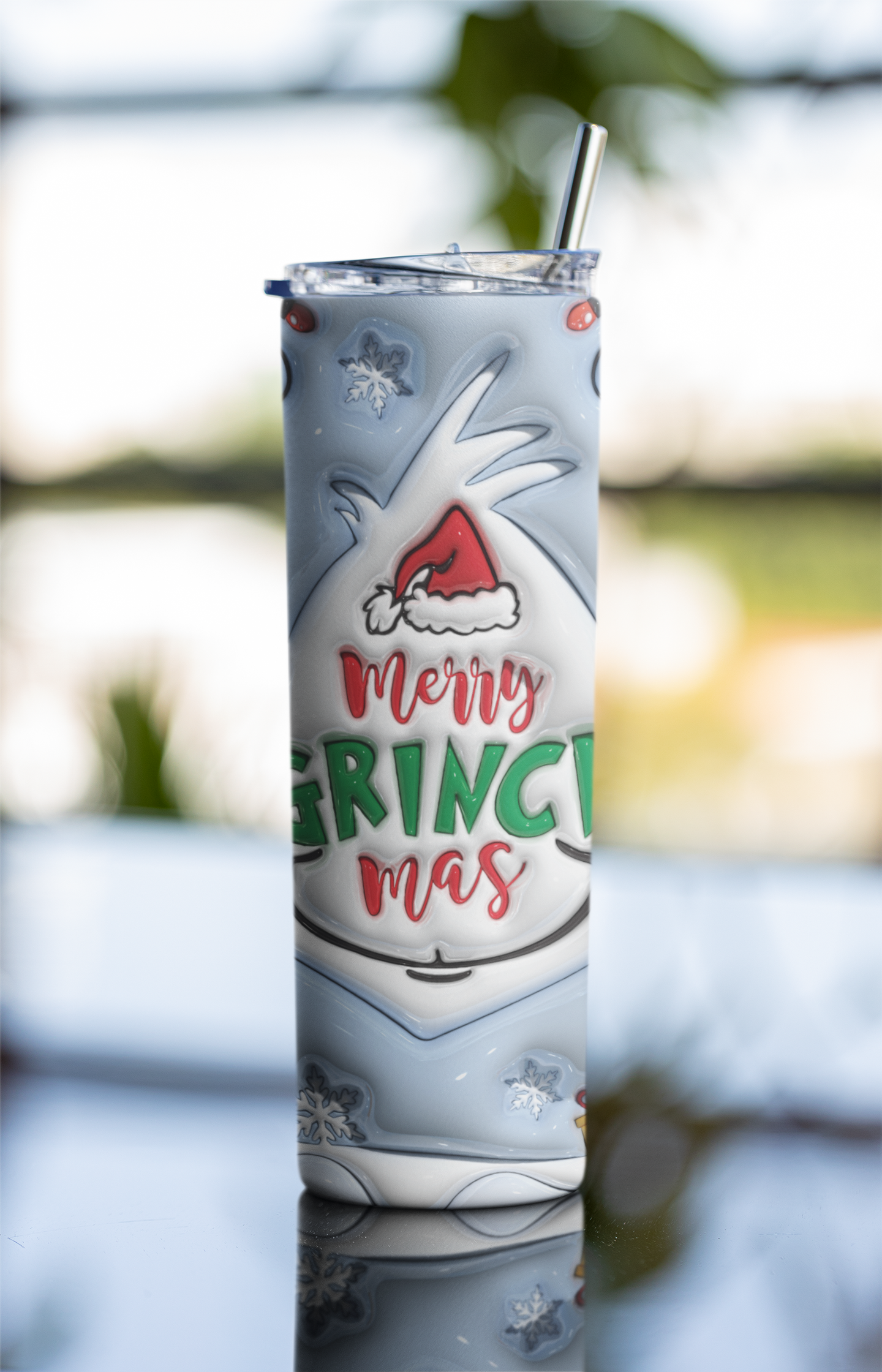 A tall insulated tumbler with a clear lid and metal straw, printed with a Grinch-themed holiday design including the text 'Merry Grinchmas' and snowflake motifs, photographed against a softly blurred outdoor background.