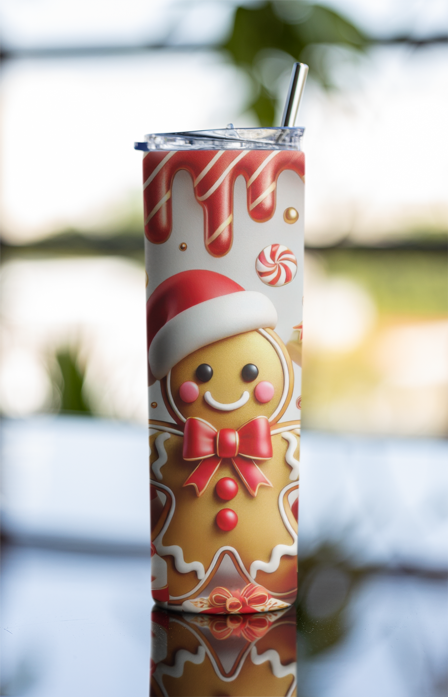 A tall insulated tumbler with a clear lid and metal straw, printed with a festive gingerbread man design in red, white, and tan, set against a softly blurred outdoor background.