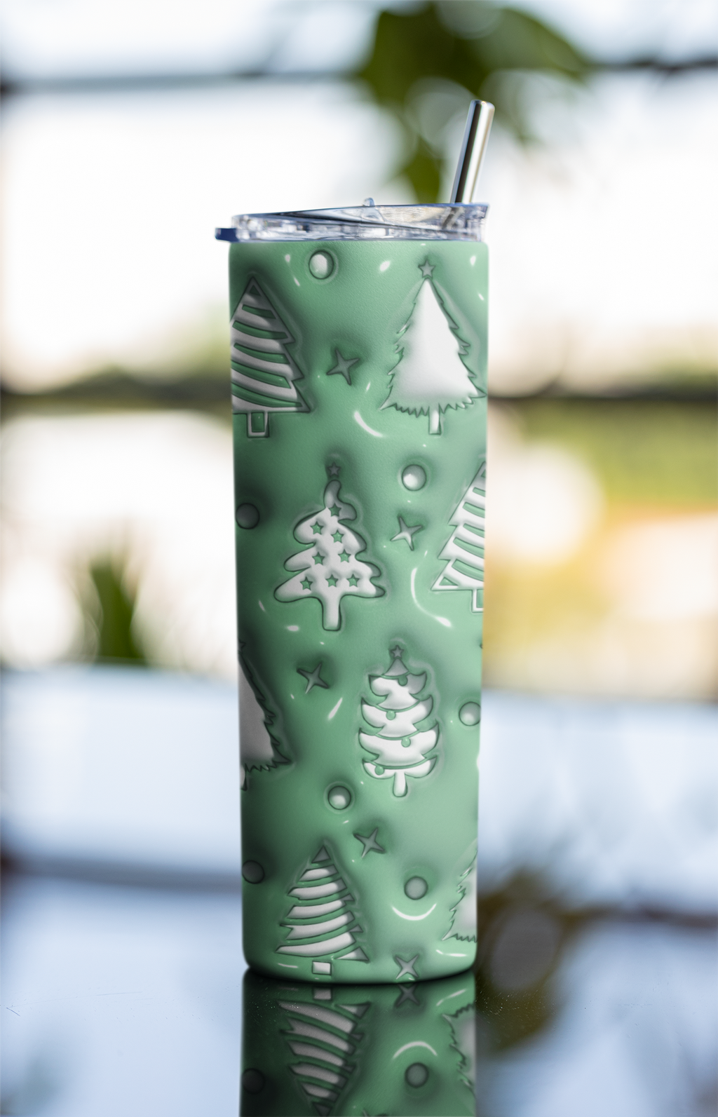 A tall insulated tumbler with a clear lid and metal straw, covered in a mint green holiday pattern of white Christmas trees, stars, and circles, set against a blurred outdoor background.