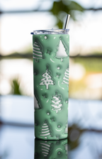 A tall insulated tumbler with a clear lid and metal straw, covered in a mint green holiday pattern of white Christmas trees, stars, and circles, set against a blurred outdoor background.