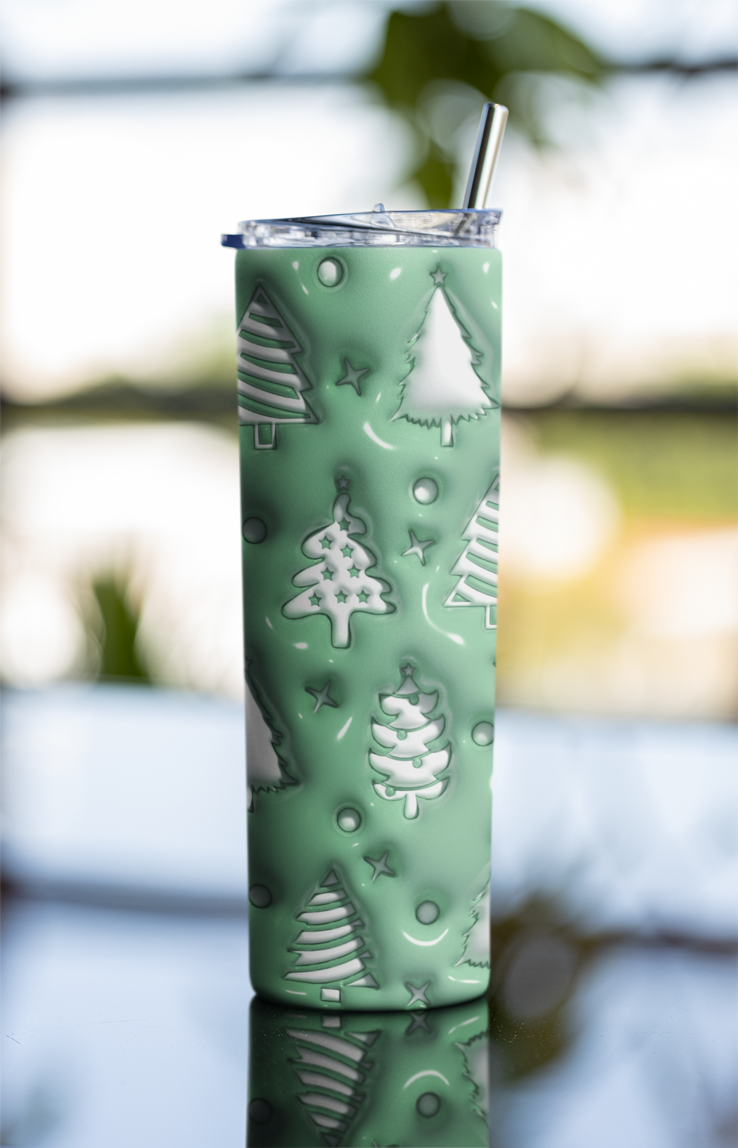 A tall insulated tumbler with a clear lid and metal straw, covered in a mint green holiday pattern of white Christmas trees, stars, and circles, set against a blurred outdoor background.