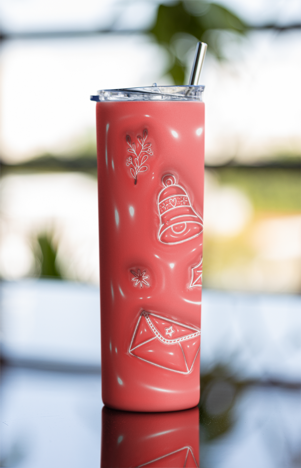 A coral pink insulated tumbler with a clear lid and metal straw, decorated with white holiday-themed embossed designs including a bell, envelope, snowflake, and pine branch. The tumbler is photographed standing upright on a reflective surface with a softly blurred outdoor background.