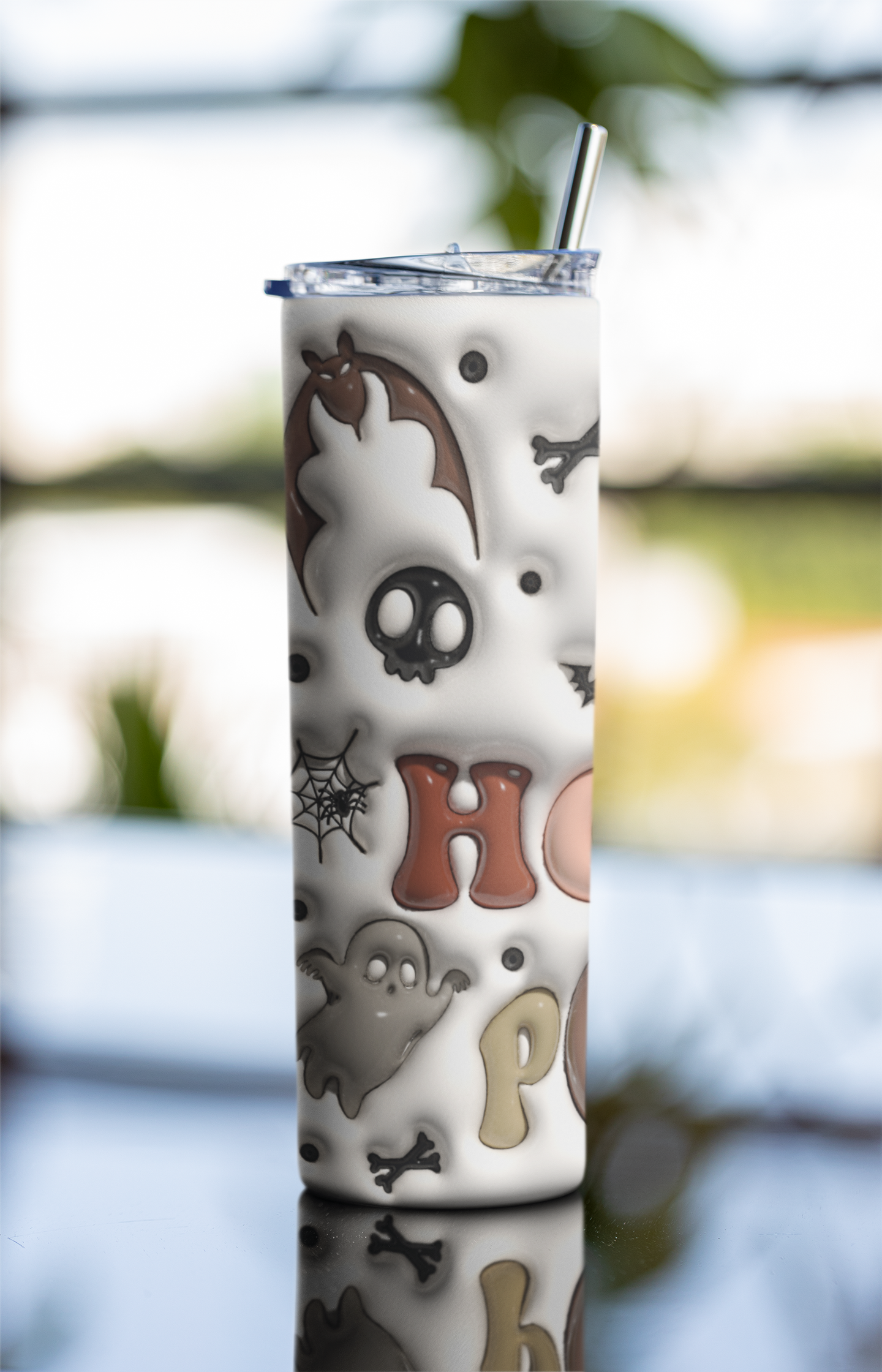 A tall white tumbler with a clear lid and metal straw, decorated with a Halloween pattern including ghosts, bats, skulls, spiderwebs, and the letters 'HO' in brown and beige. The tumbler is shown standing on a reflective surface with a blurred outdoor background.