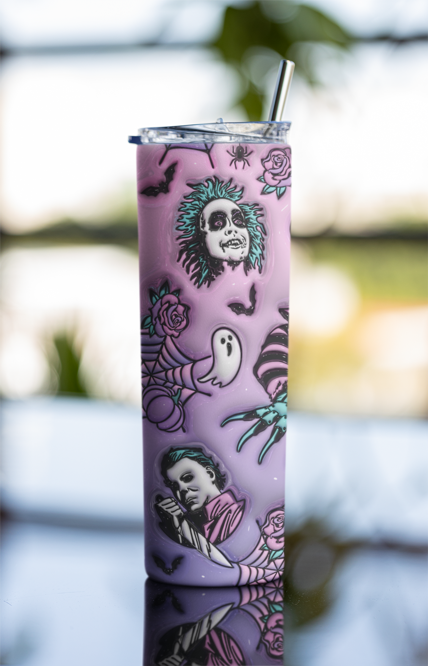 A tall lavender tumbler with a clear lid and metal straw, decorated with gothic illustrations including Beetlejuice, roses, bats, a ghost, and a spider. The tumbler is photographed on a reflective surface with a blurred outdoor background.