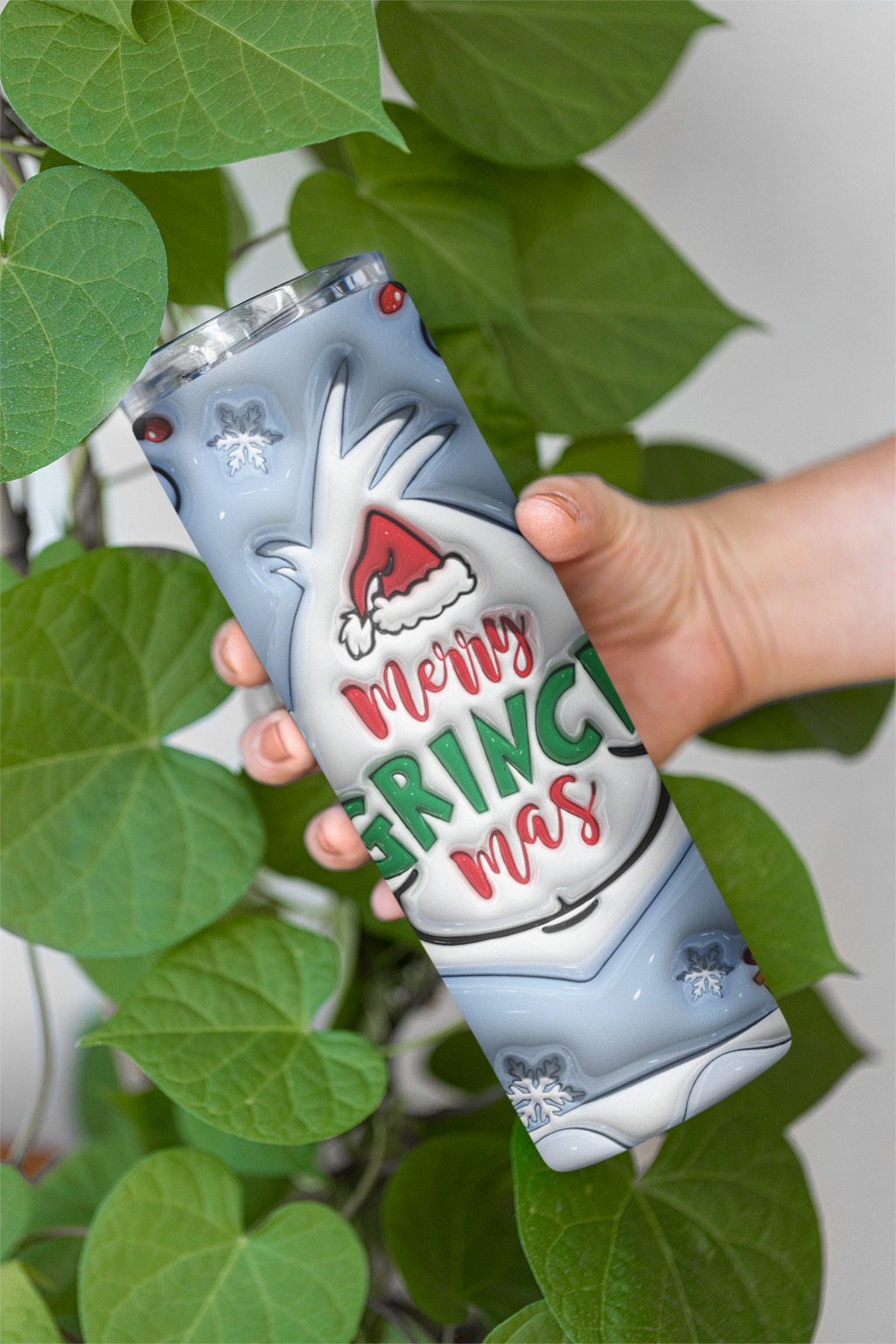 A hand holds a slim insulated tumbler with a festive 'Merry Grinch Christmas' design, surrounded by green heart-shaped leaves against a light gray background.