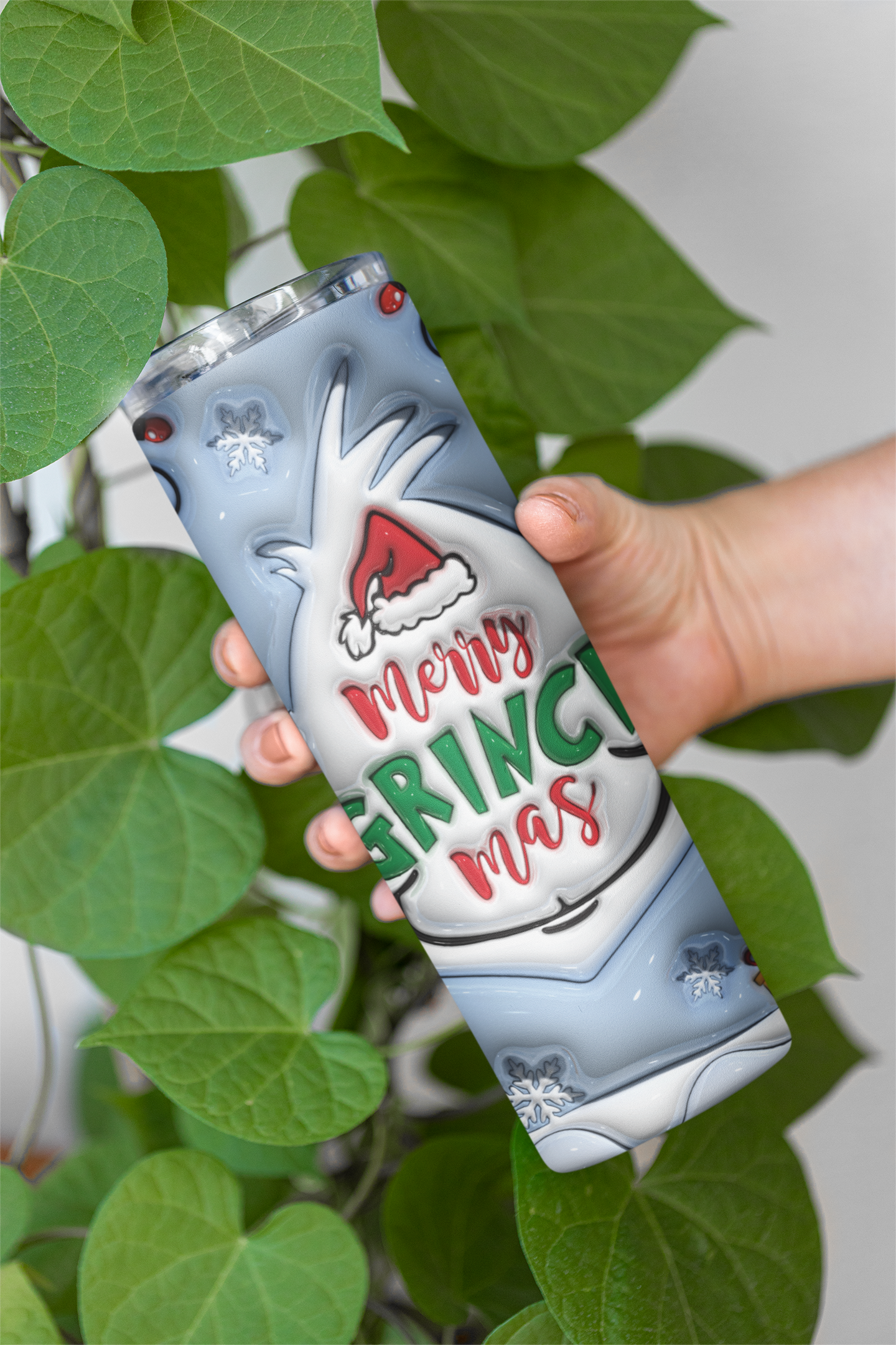 A hand holds a slim insulated tumbler with a festive 'Merry Grinch Christmas' design, surrounded by green heart-shaped leaves against a light gray background.
