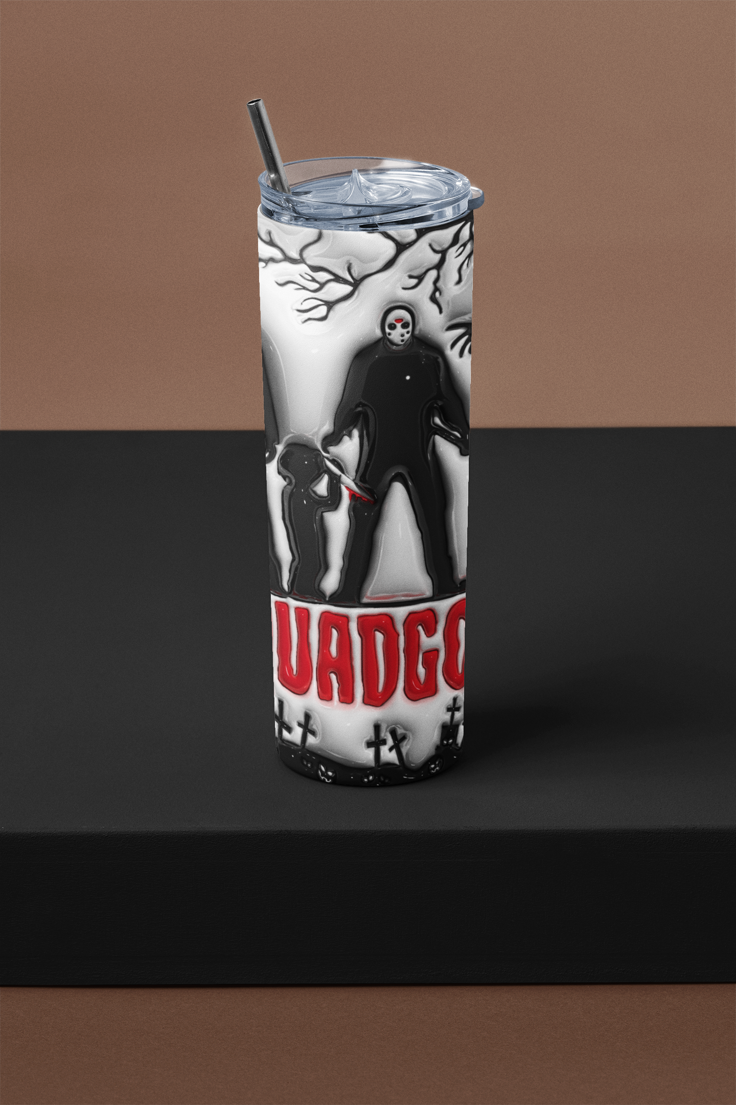 A tall insulated tumbler with a clear lid and black straw, featuring a black-and-white horror character illustration and bold red text reading 'UADGO' on a dark display surface against a brown backdrop.