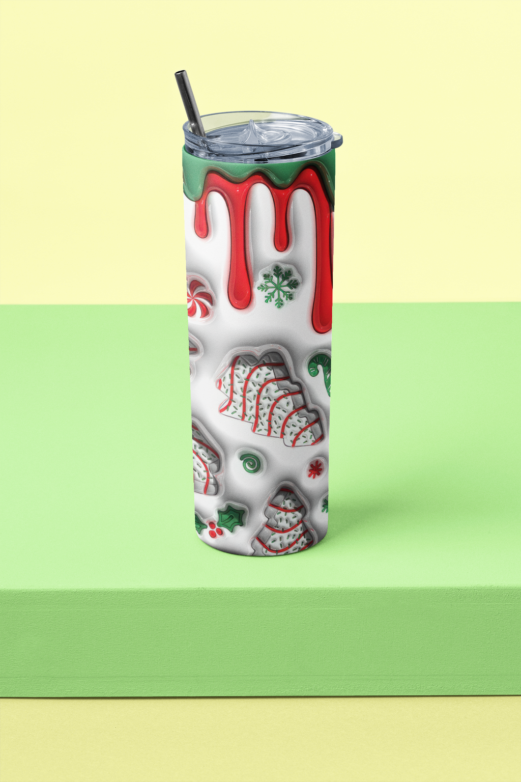 A tall insulated tumbler with a clear lid and black straw, decorated with a holiday dripping-glass pattern in red, green, and white, standing on a light green platform against a pale yellow background.