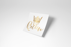 A square white sticker or card featuring a gold foil crown and the word 'Queen' in elegant gold script, shown at an angle on a light gray background with a soft shadow.