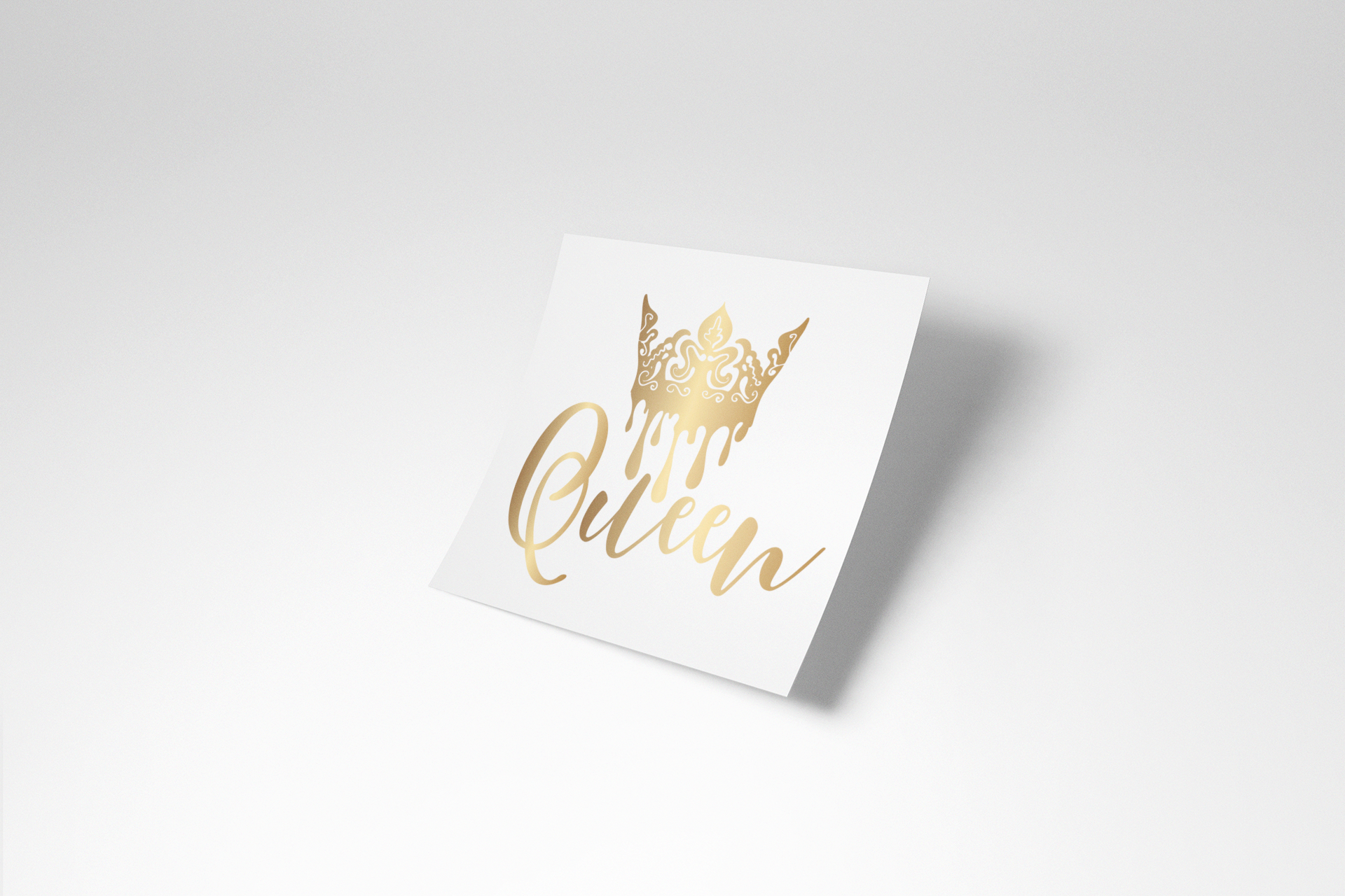 A square white sticker or card featuring a gold foil crown and the word 'Queen' in elegant gold script, shown at an angle on a light gray background with a soft shadow.