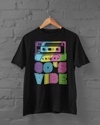 A black short-sleeve t-shirt hanging on a wooden hanger against a gray brick wall. The shirt features a colorful retro graphic of a cassette tape and the text '80'S VIBE' in neon pink, blue, yellow, and green.