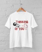 White t-shirt with red text and graphics on a hanger against a gray brick wall.