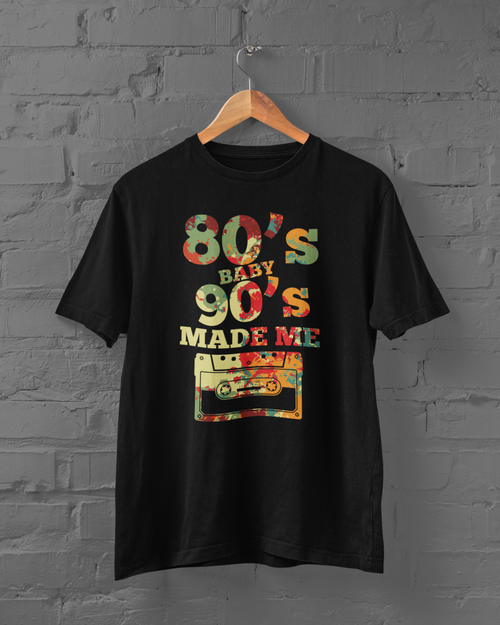 A black short-sleeve t-shirt hanging on a wooden hanger against a gray brick wall. The shirt features a colorful retro graphic with the text '80's BABY 90's MADE ME' and a cassette tape illustration.