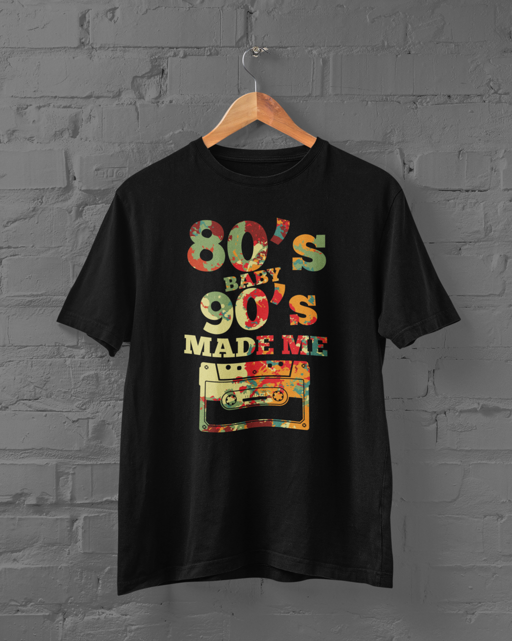 A black short-sleeve t-shirt hanging on a wooden hanger against a gray brick wall. The shirt features a colorful retro graphic with the text '80's BABY 90's MADE ME' and a cassette tape illustration.