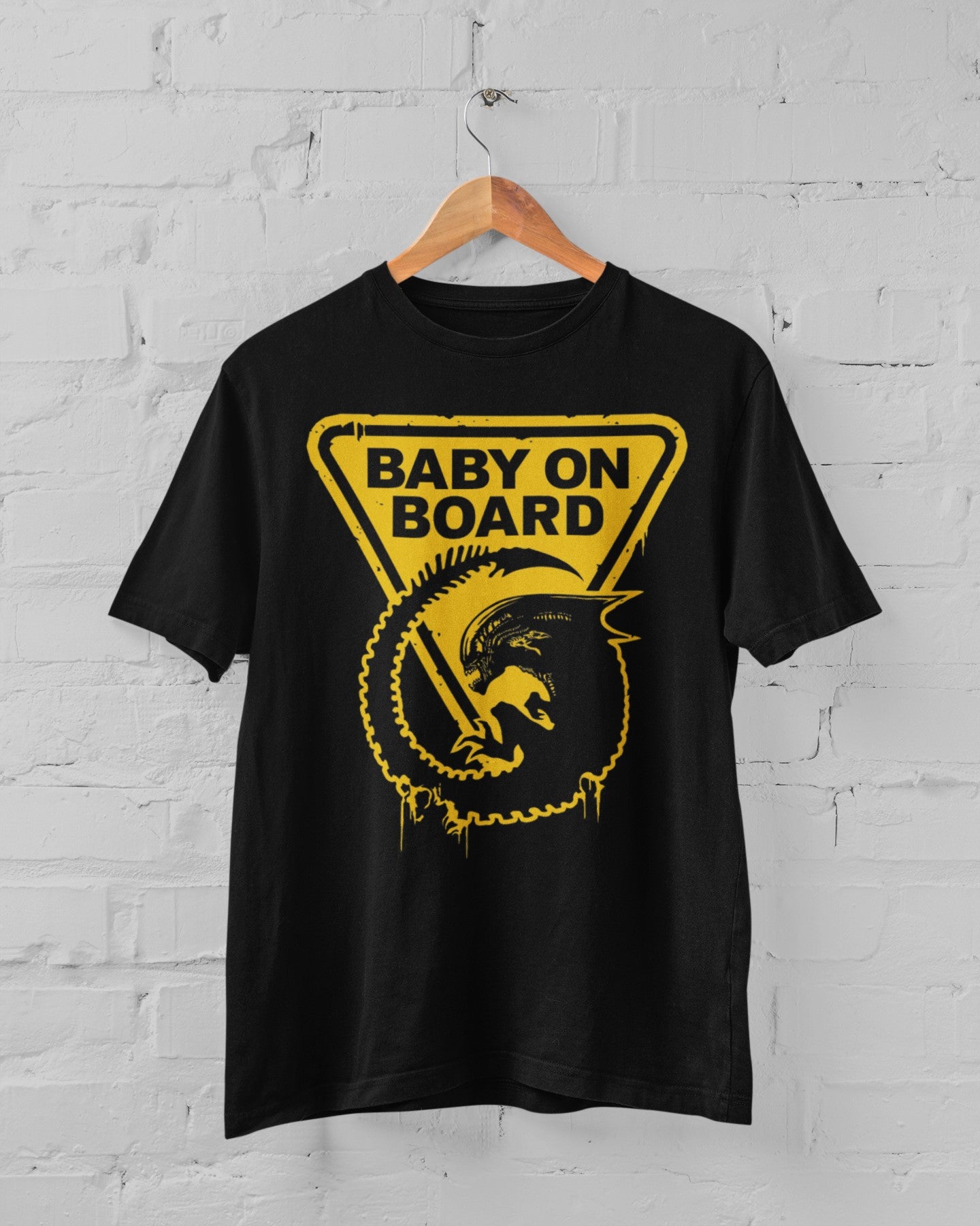 Black t-shirt with yellow 'Baby on Board' graphic on a hanger against a white brick wall.