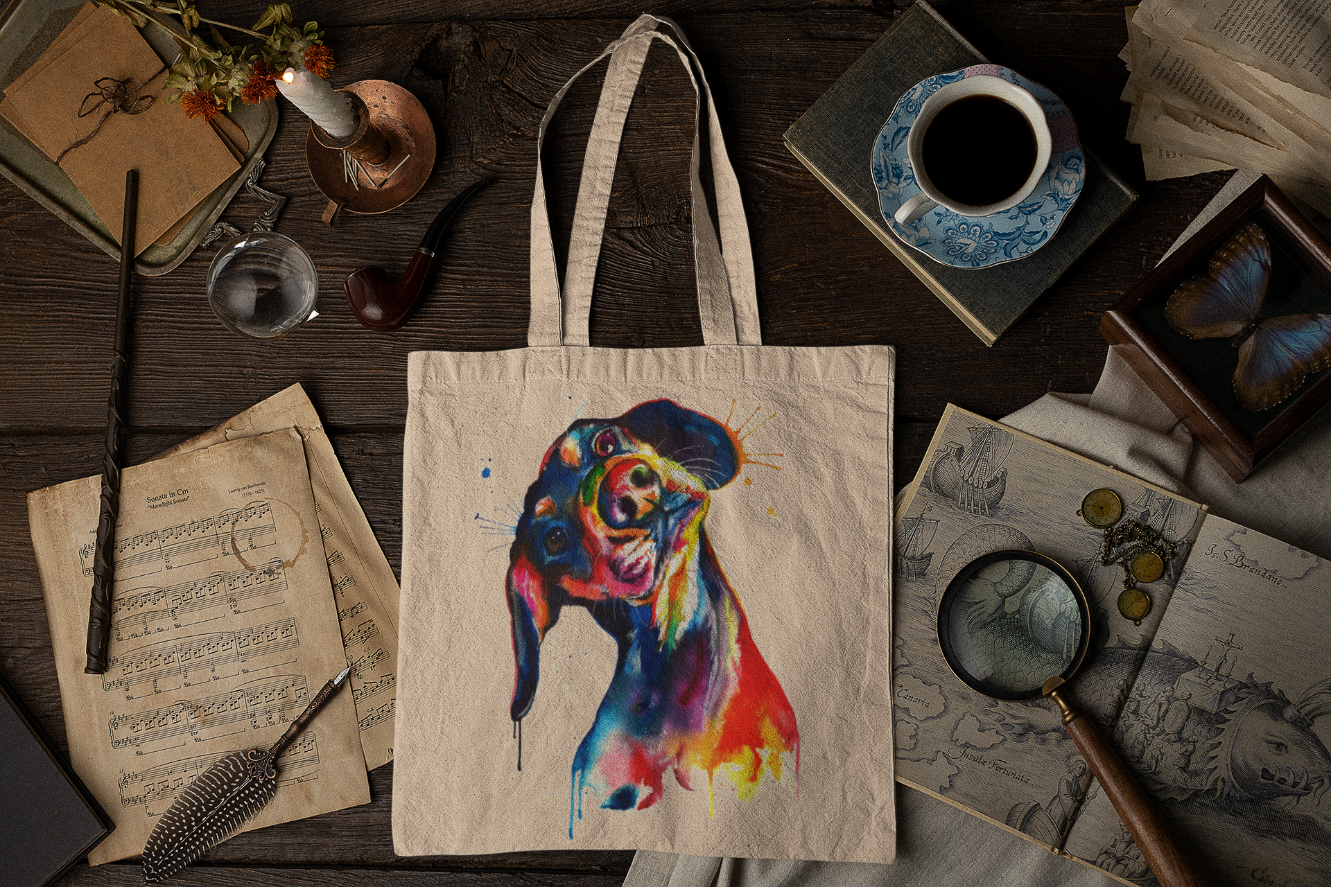 A canvas tote bag with a colorful watercolor-style dog portrait, displayed in a styled flat-lay on a dark wooden table surrounded by vintage books, a cup of coffee, a magnifying glass, and other antique props.