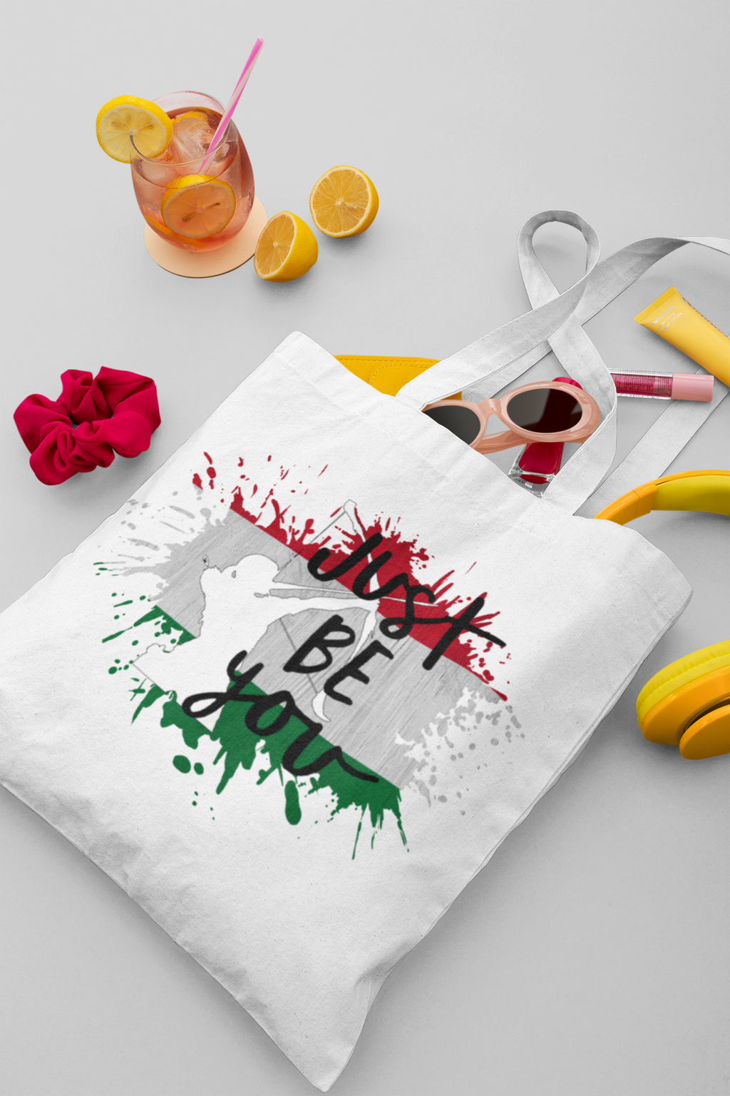Tote bag with colorful design and text, surrounded by a drink, fruit, and accessories on a light gray background.