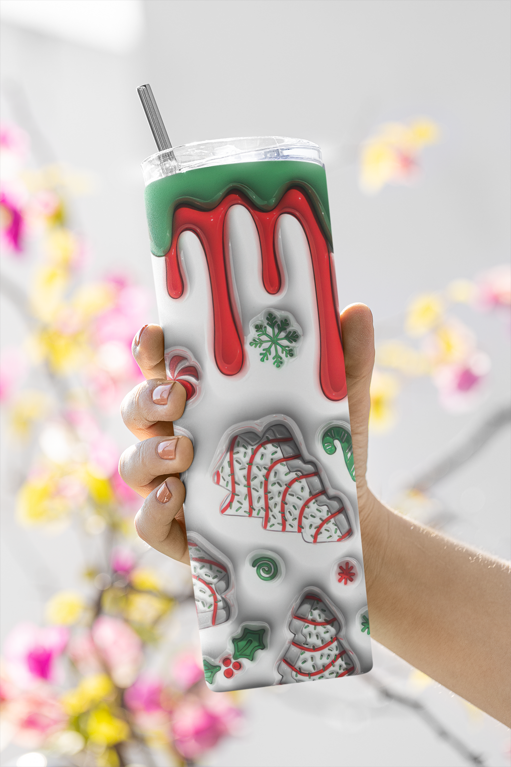 A hand holding a clear plastic tumbler with a metal straw. The tumbler has a white background decorated with red dripping icing, green icing, and various holiday cookie illustrations.