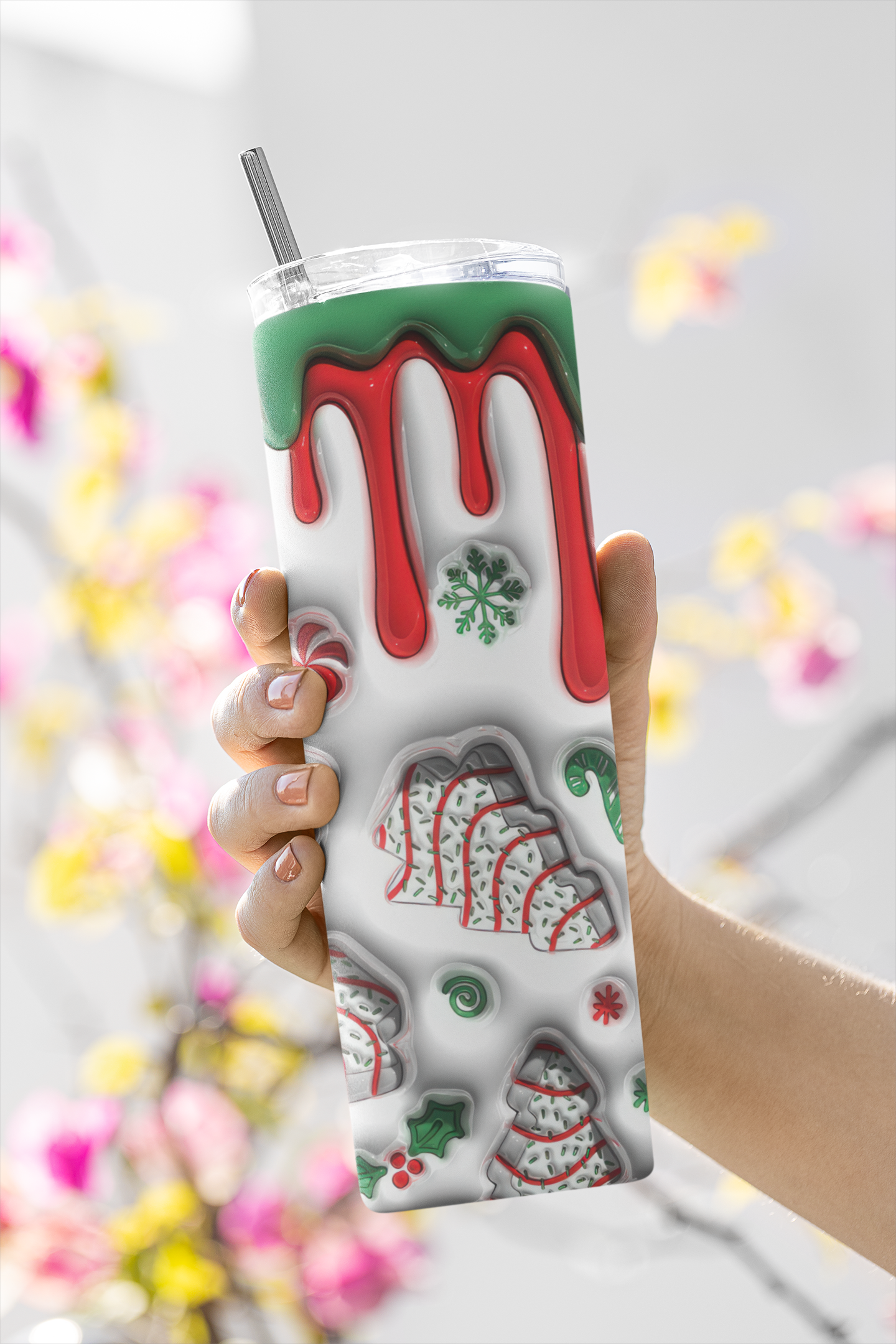 A hand holding a clear plastic tumbler with a metal straw. The tumbler has a white background decorated with red dripping icing, green icing, and various holiday cookie illustrations.