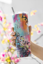 Colorful tumbler with artistic design held by a hand against a blurred floral background