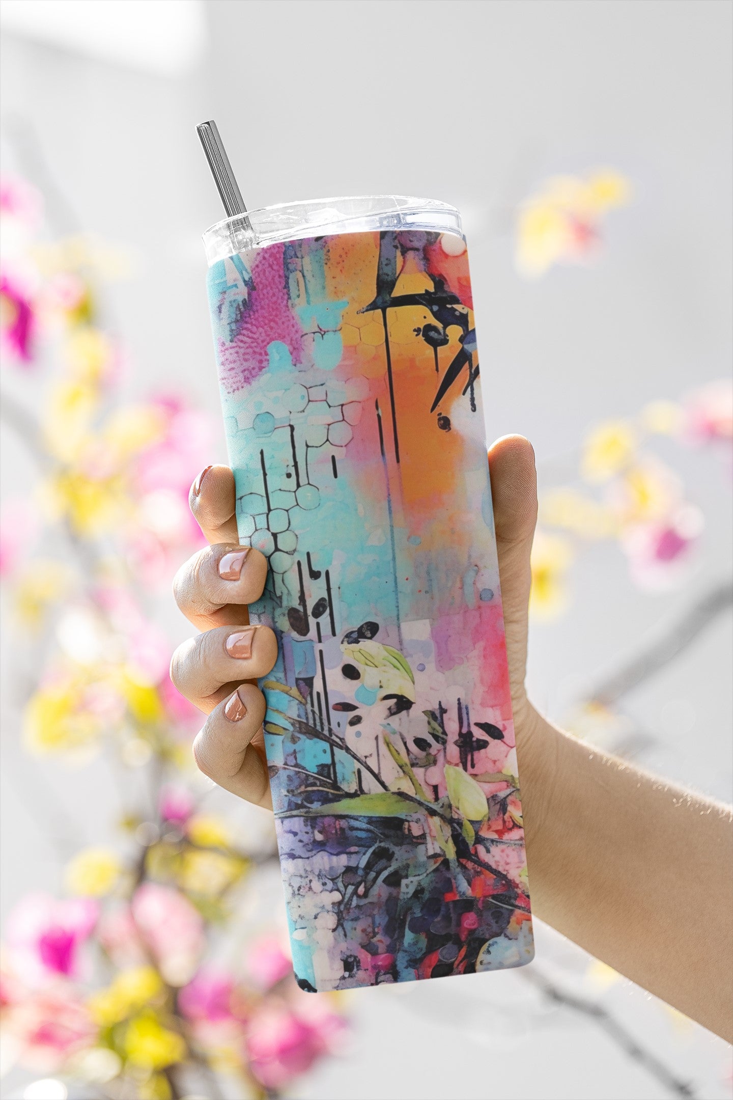 Colorful tumbler with artistic design held by a hand against a blurred floral background