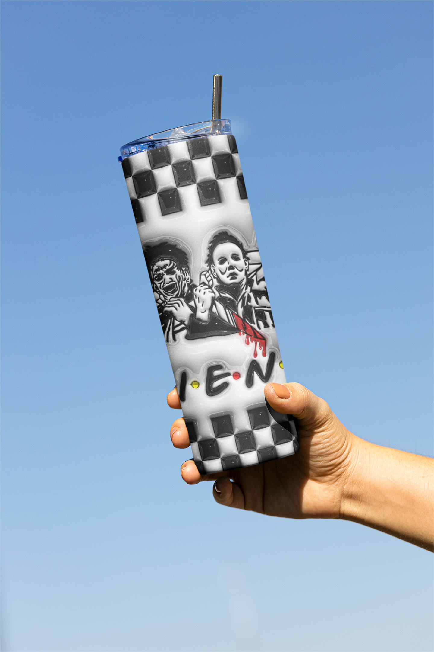 A hand holding a tall clear tumbler with a black-and-white checkerboard pattern, black-and-white graphic artwork, and a metal straw against a clear blue sky.