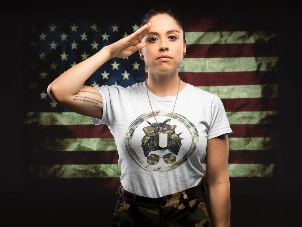 A woman saluting in front of a distressed American flag background, wearing a light gray t-shirt with a military-themed graphic of a dog wearing a dog collar and sunglasses, paired with camouflage pants.