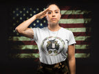 A woman saluting in front of a distressed American flag background, wearing a light gray t-shirt with a military-themed graphic of a dog wearing a dog collar and sunglasses, paired with camouflage pants.