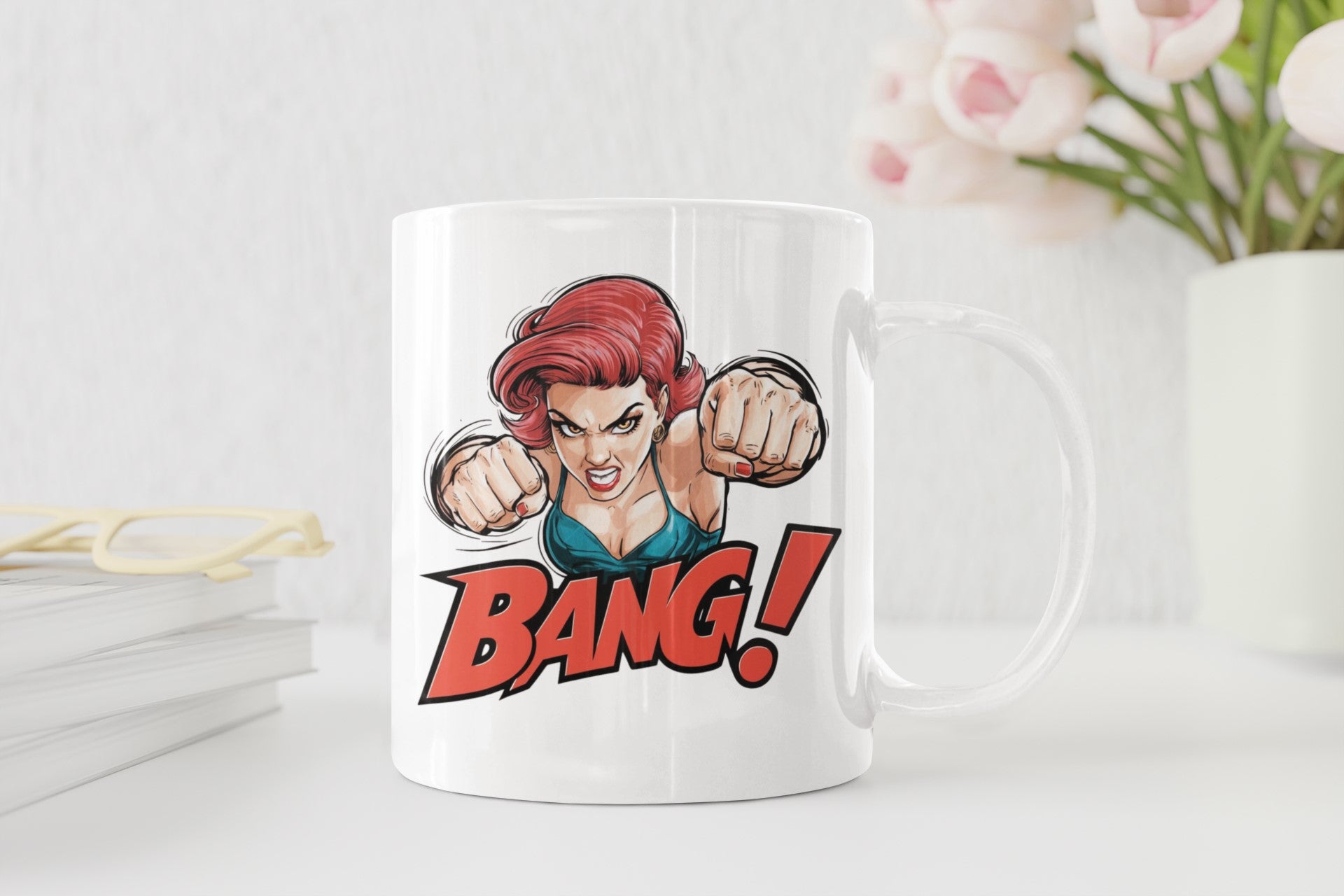 A white ceramic mug with a comic-style graphic of a red-haired woman punching forward above the word 'BANG!' in bold red letters, shown on a light tabletop with books, glasses, and flowers in the background.
