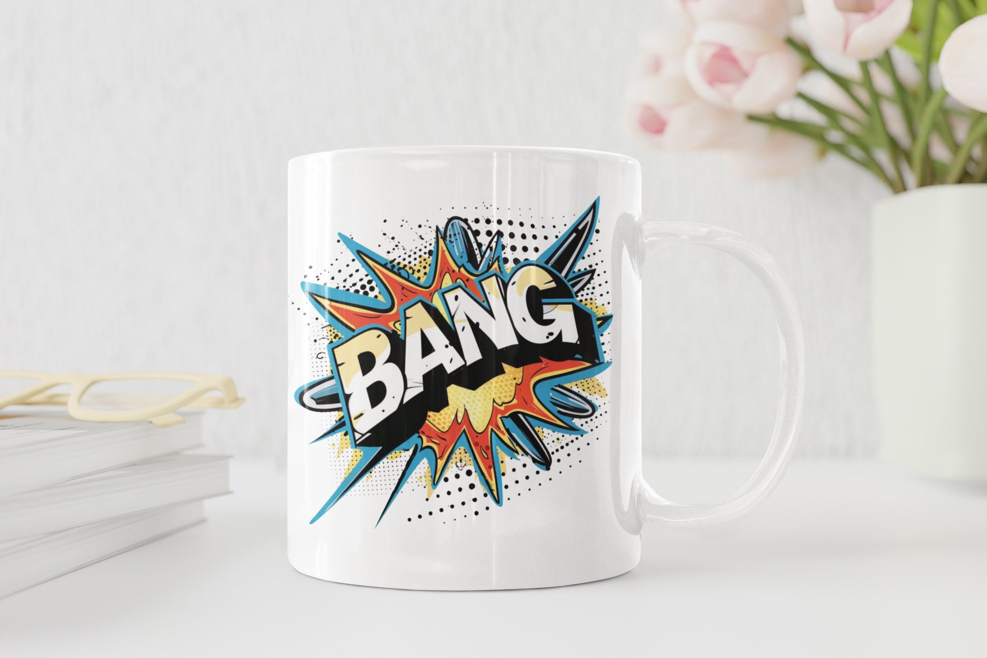 A white ceramic mug with a colorful comic-style 'BANG' speech bubble graphic, set on a white surface with a blurred background of books, glasses, and pink flowers.