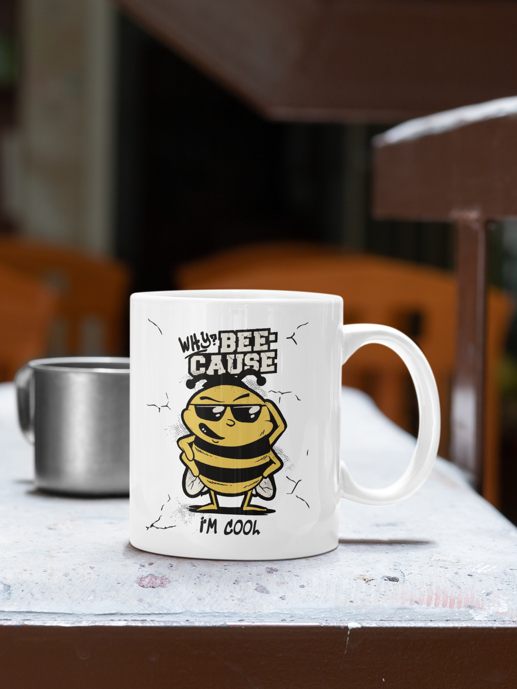 A white ceramic coffee mug with a cartoon bee graphic and the text 'WHY? I'M BEE-CAUSE I'M COOK.' sitting on a table in a cafe setting.