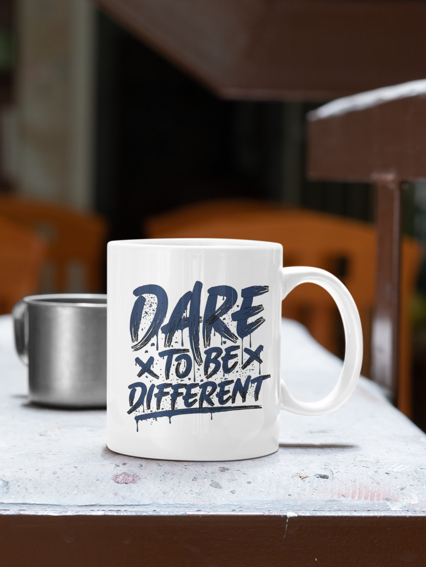 A white ceramic coffee mug with the text 'DARE TO BE DIFFERENT' printed in a dark navy blue graffiti-style font, sitting on a weathered gray table in an outdoor cafe setting.