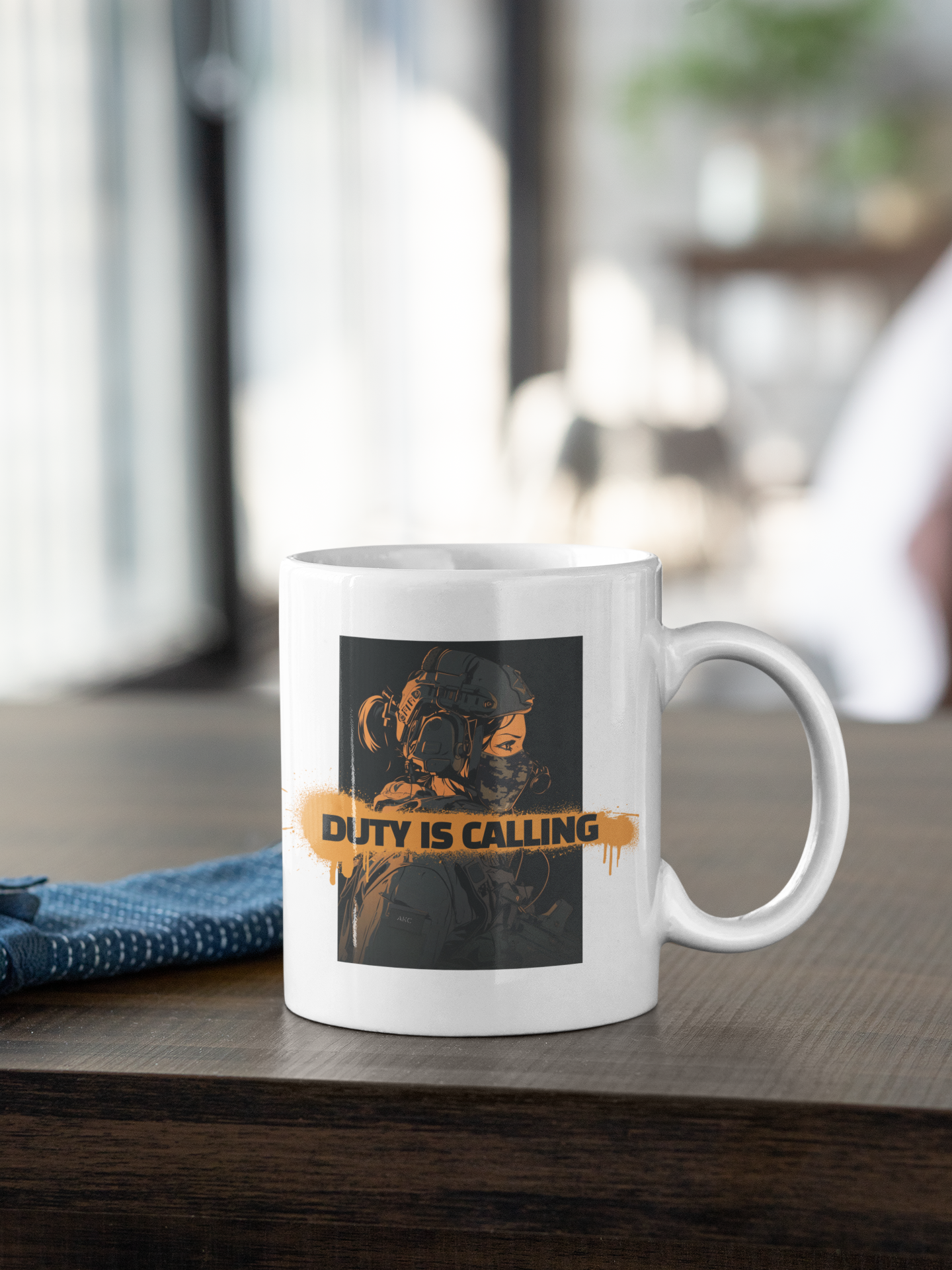 A white ceramic coffee mug with a graphic print of a soldier in a gas mask and the text 'DUTY IS CALLING' in orange, sitting on a dark wooden table in a blurred indoor setting.