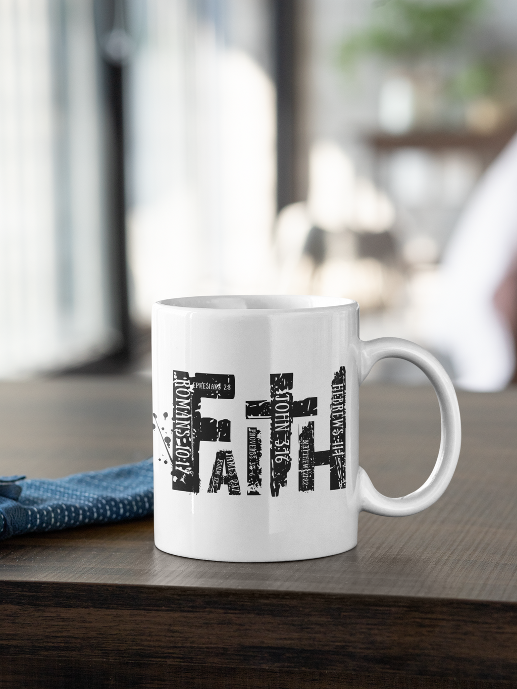 A white ceramic coffee mug with a black typographic cross design spelling out 'FAITH' with smaller words like 'JUDICIOUS FAITH' and 'HOPEFUL FAITH' integrated into the letters. The mug is sitting on a dark wooden table next to a blue textured cloth, with a softly blurred indoor background.