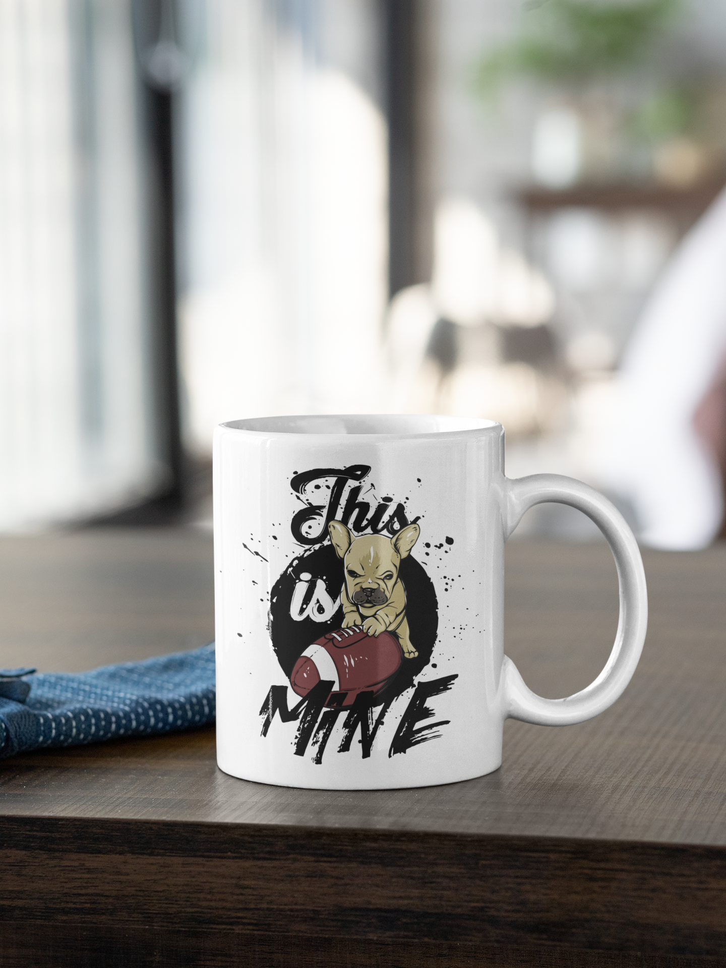 A white ceramic coffee mug with a graphic print of a French bulldog holding a football and the text 'This is MINE', sitting on a dark wooden table in a blurred indoor setting.