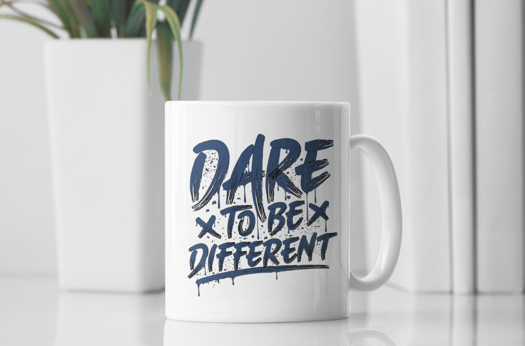 A white ceramic mug with the text 'DARE TO BE DIFFERENT' printed in dark navy blue, set on a reflective white surface with a blurred white plant in the background.