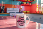 A white ceramic mug with a pink flamingo graphic and the text 'don't make me put my foot down' sits on a pink-tinted kitchen counter with a blurred colorful kitchen in the background.