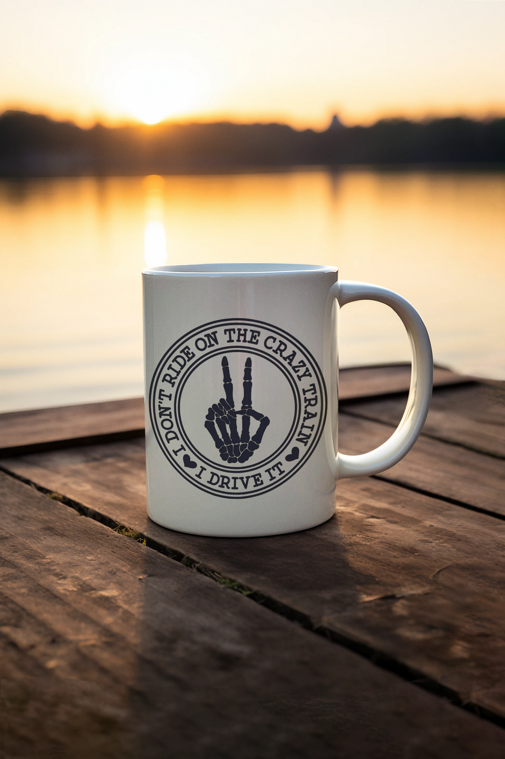 A white ceramic coffee mug with a black circular graphic sitting on a wooden dock at sunset with a lake in the background. The graphic features a skeleton hand making a middle finger gesture with the text 'DON'T RIDE ON THE CRAZY TRAIL I DRIVE NIN'.