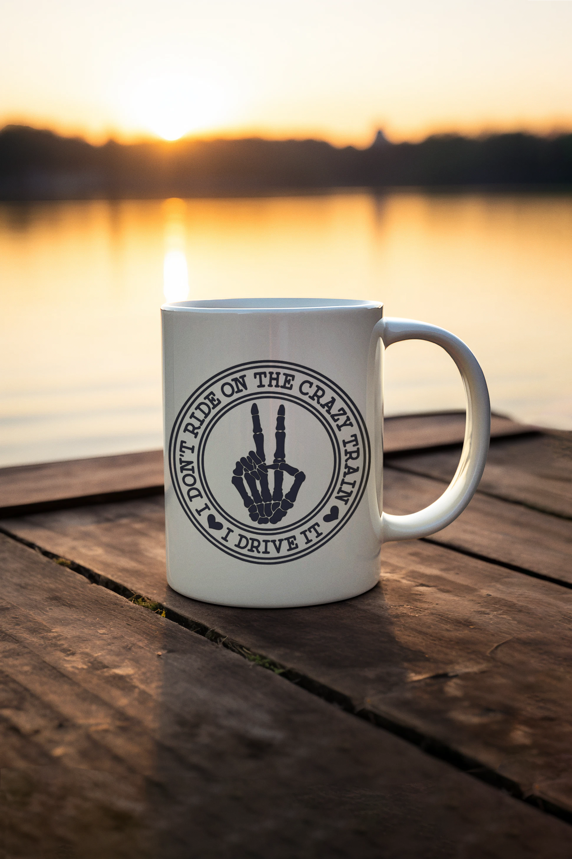 A white ceramic coffee mug with a black circular graphic sitting on a wooden dock at sunset with a lake in the background. The graphic features a skeleton hand making a middle finger gesture with the text 'DON'T RIDE ON THE CRAZY TRAIL I DRIVE NIN'.