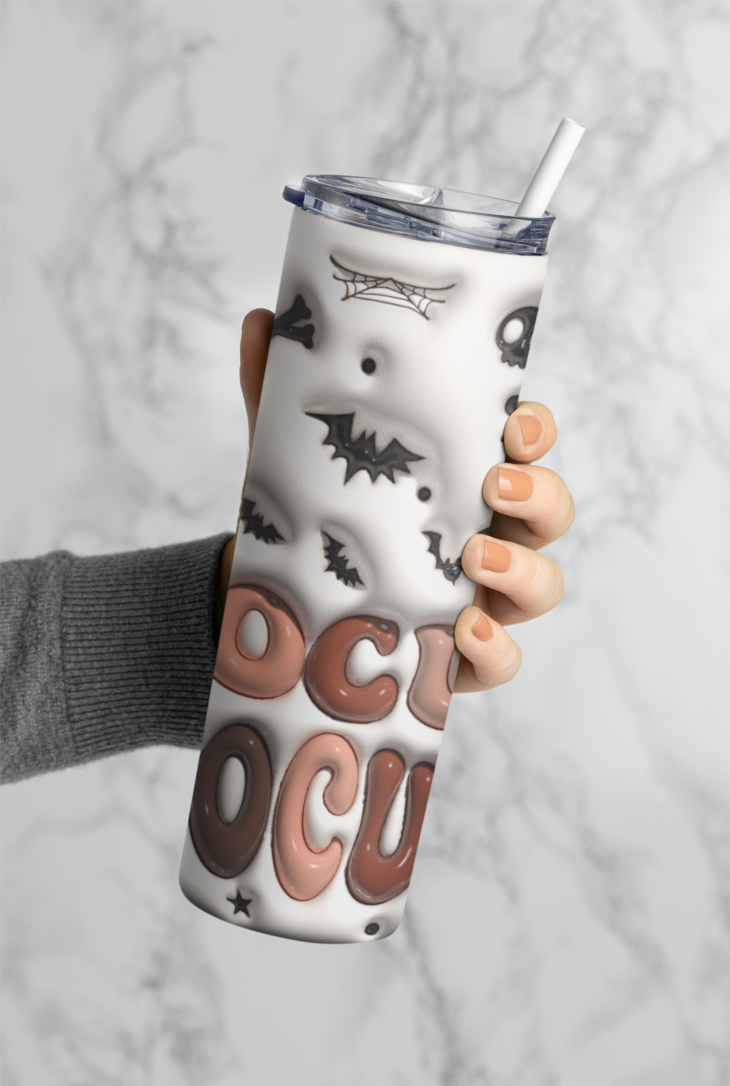 A hand holding a white tumbler with a clear lid and straw, decorated with black bat and spider graphics and large 'OCCU' lettering in brown and pink tones, against a light marble background.