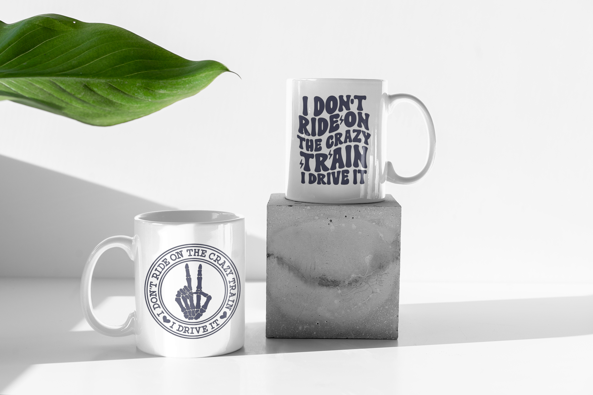 Two white ceramic mugs with black printed text and a hand gesture design, styled on a white surface with a concrete block and a green leaf in the background.