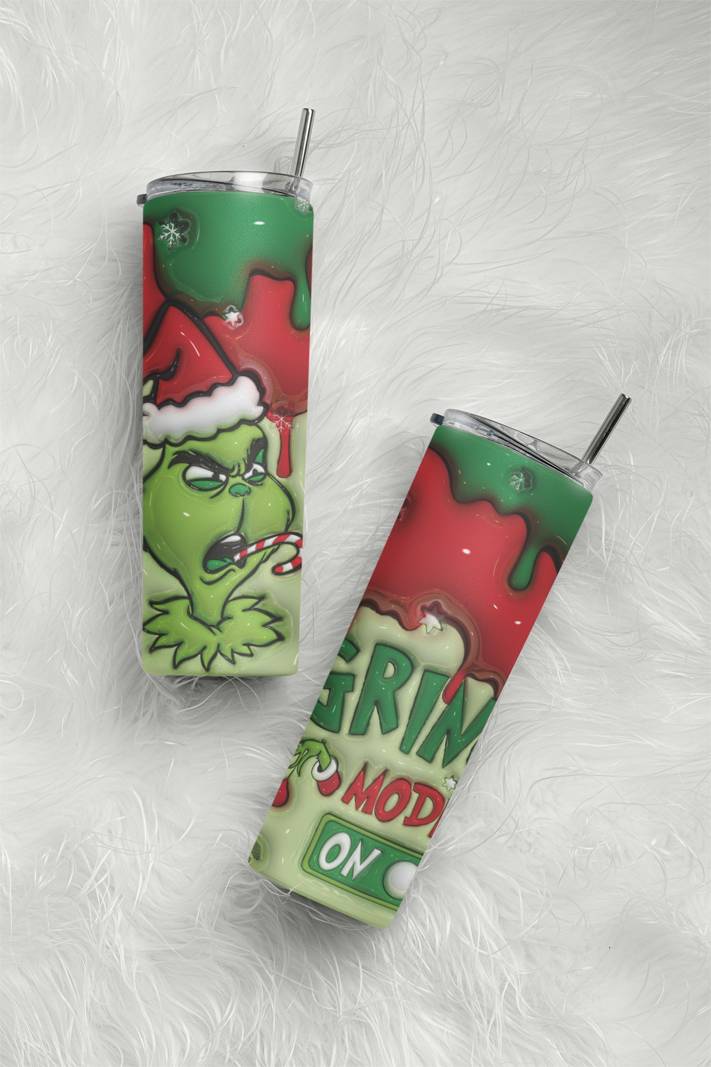 Two slim insulated tumblers with clear lids and metal straws, printed with a Grinch-themed Christmas design, placed on a white faux-fur surface. One tumbler shows the Grinch wearing a Santa hat, and the other shows partial holiday text and graphics.