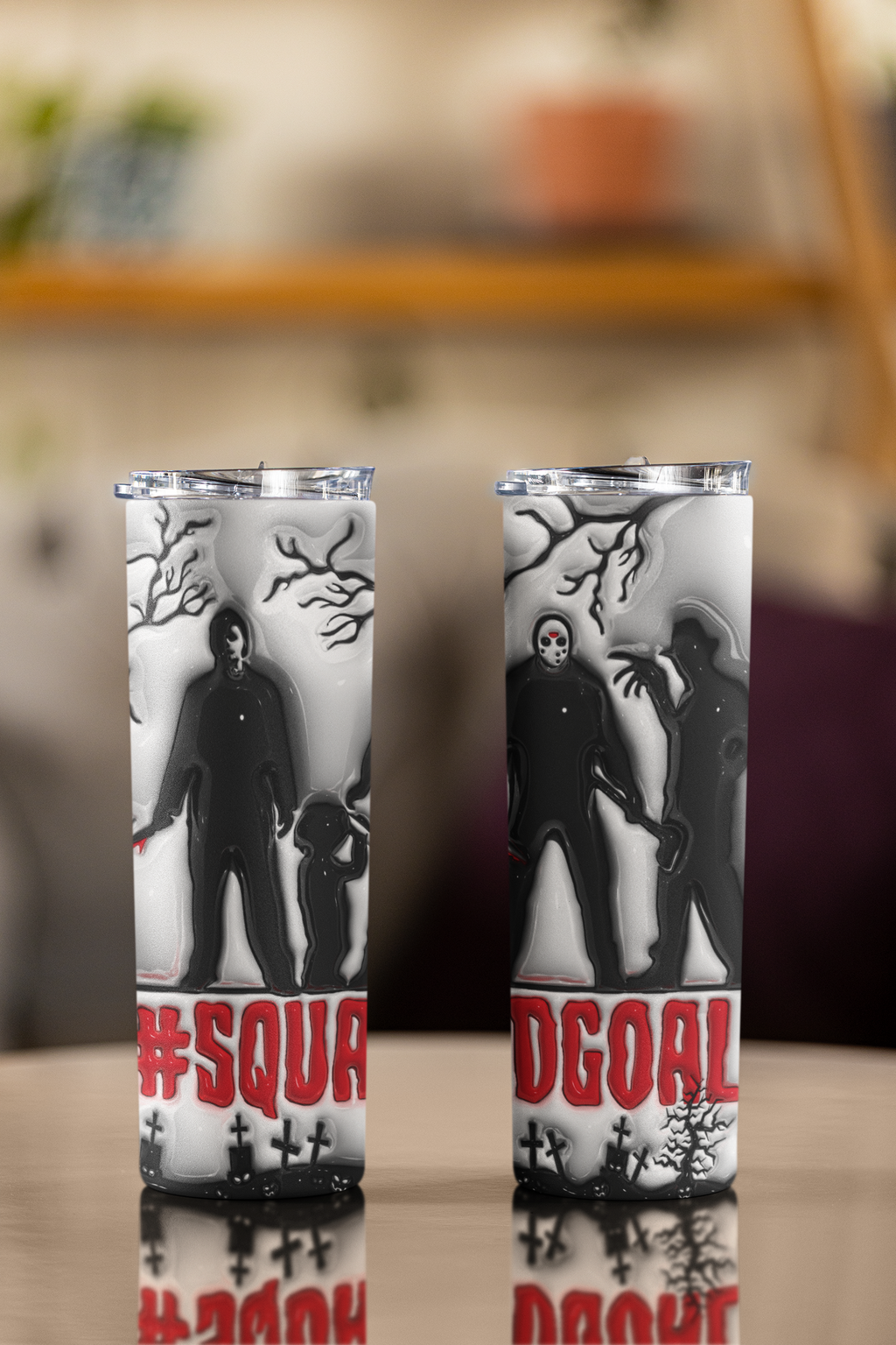 Two tall insulated tumblers with clear lids, featuring a black and white horror-themed graphic of a masked figure and zombie-like silhouette with red text at the bottom, displayed on a reflective tabletop against a blurred indoor background.