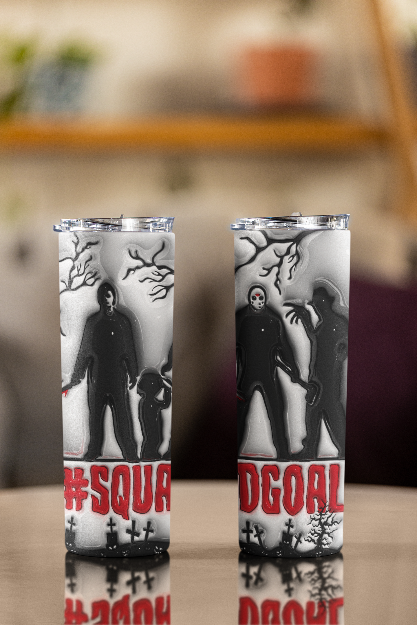 Two tall insulated tumblers with clear lids, featuring a black and white horror-themed graphic of a masked figure and zombie-like silhouette with red text at the bottom, displayed on a reflective tabletop against a blurred indoor background.