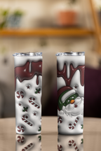Two tall insulated tumblers with clear lids, shown side by side on a tabletop. The tumblers have a white holiday design featuring dark brown chocolate drizzle, candy canes, peppermint stars, and a Santa Claus face on the right tumbler.