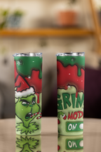 Two slim stainless steel tumblers with clear lids, featuring a Grinch-themed Christmas design with green and red colors, standing on a reflective tabletop against a blurred indoor background.