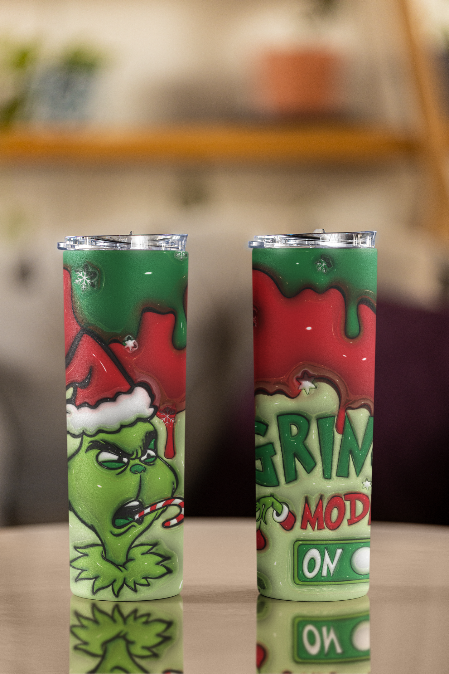 Two slim stainless steel tumblers with clear lids, featuring a Grinch-themed Christmas design with green and red colors, standing on a reflective tabletop against a blurred indoor background.