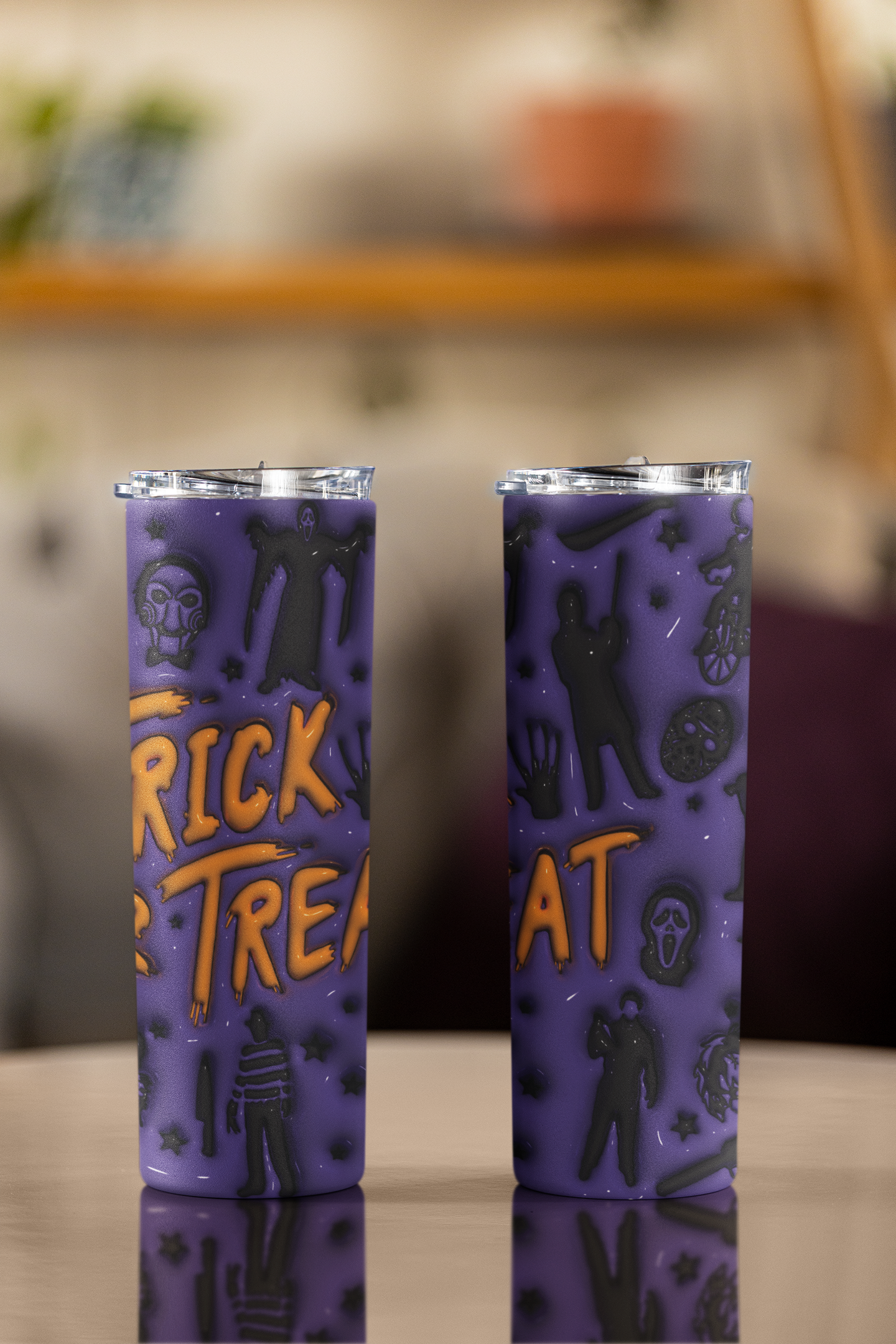 Two purple insulated tumblers with a Halloween theme, featuring black silhouettes of ghosts, zombies, and a ghost face, with orange 'TRICK OR TREAT' lettering on the front, displayed on a reflective surface with a blurred indoor background.